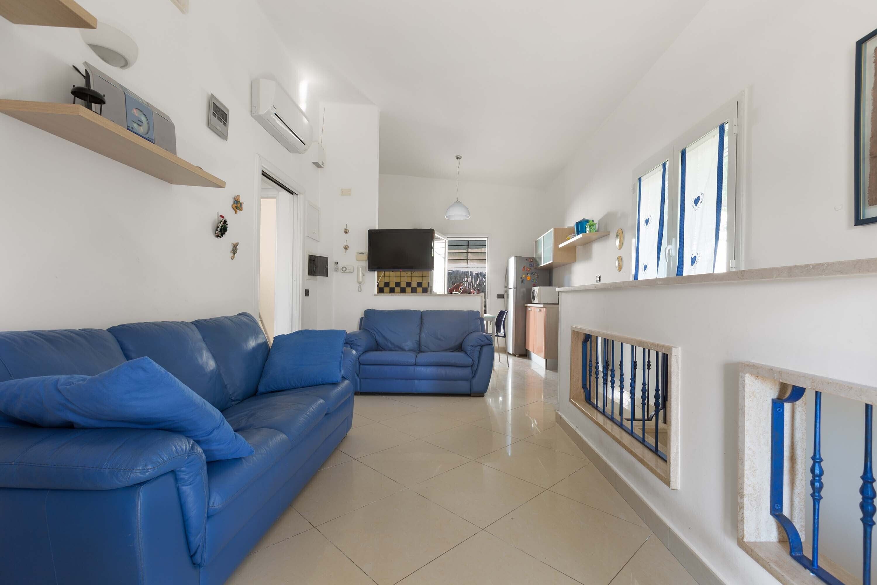 A modern living room with two blue sofas, a kitchen area with a tiled backsplash, and a hallway with a blue railing.
