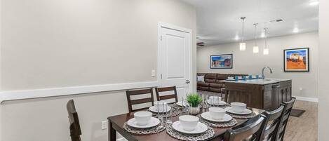 A dining area with a wooden table set for four, surrounded by chairs. There is a kitchen area in the background with a brown countertop and a sink.