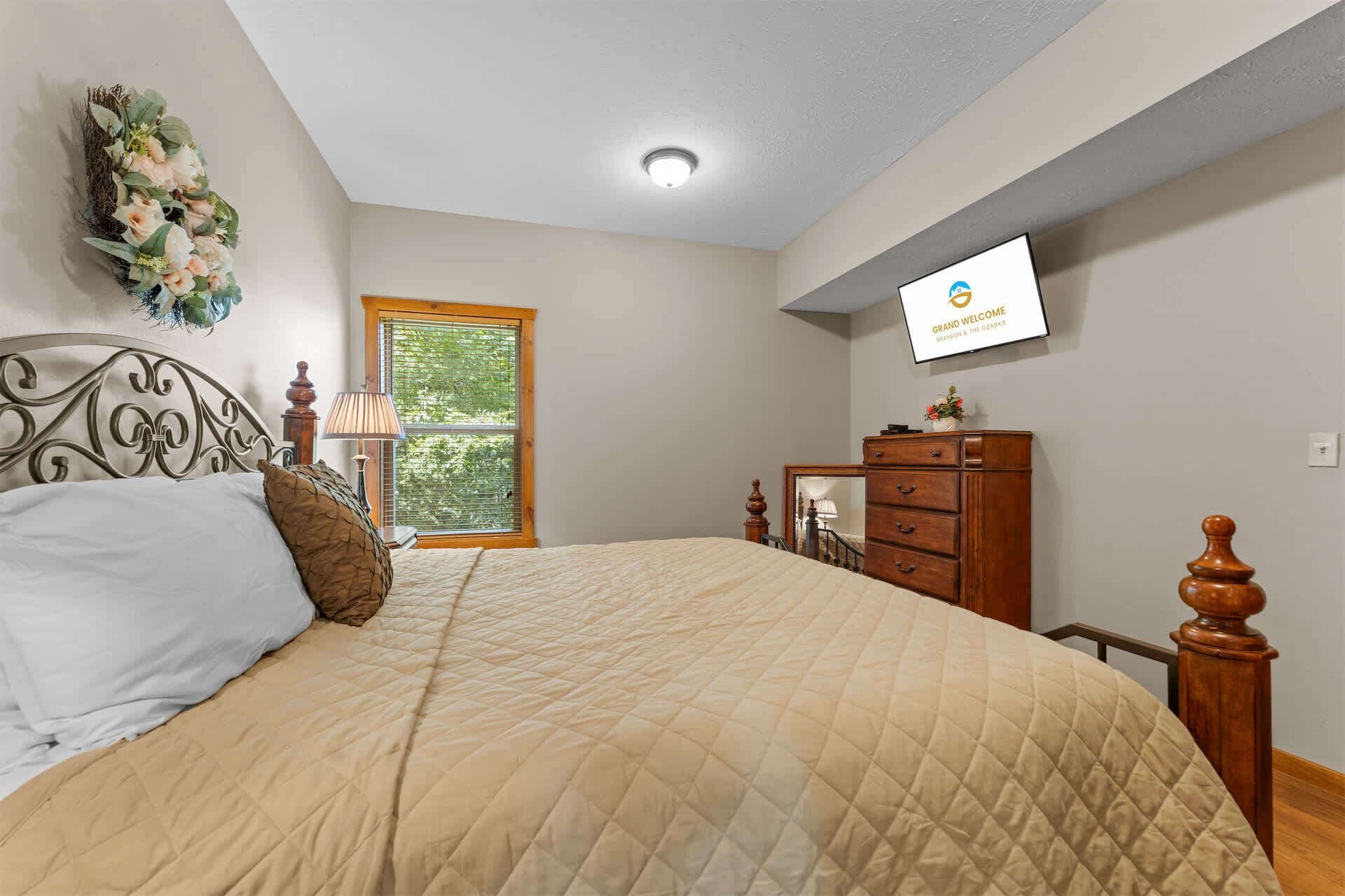 Un dormitorio con cama, cómoda, espejo y televisor mostrando 'Grand Welcome'.
