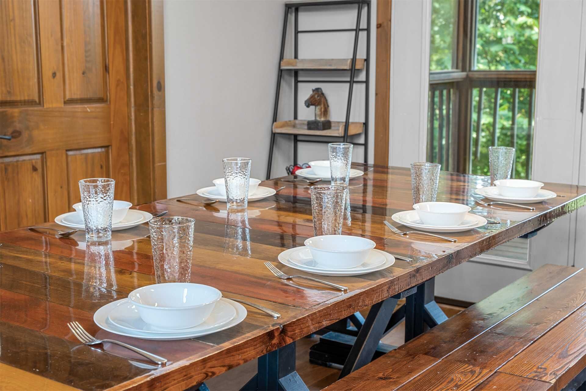 Mesa de comedor con platos blancos, tazones y vasos transparentes, con un banco de madera y una escalera al fondo.