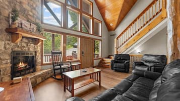 Amplia sala de estar con chimenea de piedra, mobiliario de cuero, ventanas grandes y una escalera de madera.