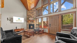A spacious living room with a stone fireplace, wooden ceiling, and large windows. - Kemper's Cabin by Silver Dollar City - Ideally Located (Branson)