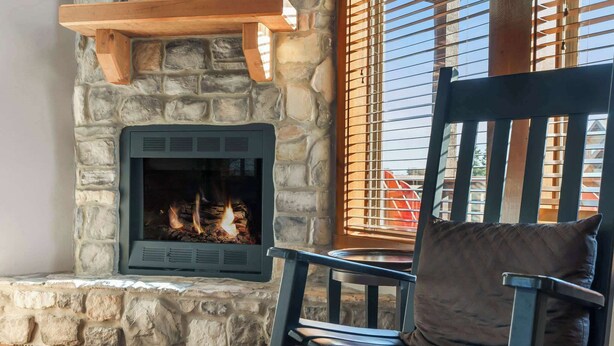 A stone fireplace with a fire, a wooden shelf above it, and a chair with a cushion in front. - Kemper's Cabin by Silver Dollar City - Ideally Located (Branson)