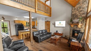 A spacious living area with a stone fireplace, a black leather sofa, a wooden dining table, and a kitchen with wooden cabinets. - Kemper's Cabin by Silver Dollar City - Ideally Located (Branson)