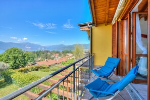 A balcony with two blue chairs, a wooden railing, and a view of a town and mountains. - La Brezza Del Lago Maggiore: Apartment With Terrace and View (Laveno Mombello)