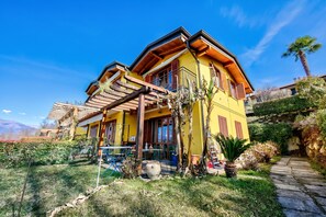 A yellow building with a wooden deck, a stone pathway, and a garden with various plants. - La Brezza Del Lago Maggiore: Apartment With Terrace and View (Laveno Mombello)