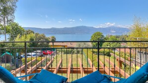 A balcony with a view of a lake and mountains, wooden deck, and a red car in the background. - La Brezza Del Lago Maggiore: Apartment With Terrace and View (Laveno Mombello)