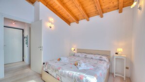A bedroom with a wooden ceiling, a bed with floral bedding, bedside lamps, and a white nightstand. - La Brezza Del Lago Maggiore: Apartment With Terrace and View (Laveno Mombello)