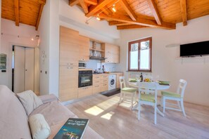 A modern kitchen with wooden beams, a dining area with white chairs, and a sofa with a book. - La Brezza Del Lago Maggiore: Apartment With Terrace and View (Laveno Mombello)