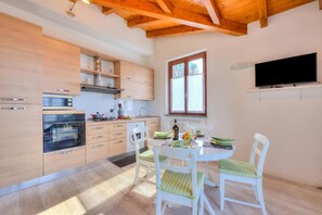 A modern kitchen with wooden cabinets, a built-in oven, and a dining area with white chairs and a table. - La Brezza Del Lago Maggiore: Apartment With Terrace and View (Laveno Mombello)