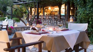 An outdoor dining area with a table set for two, surrounded by chairs. There is a pergola structure in the background with a bar area visible.
