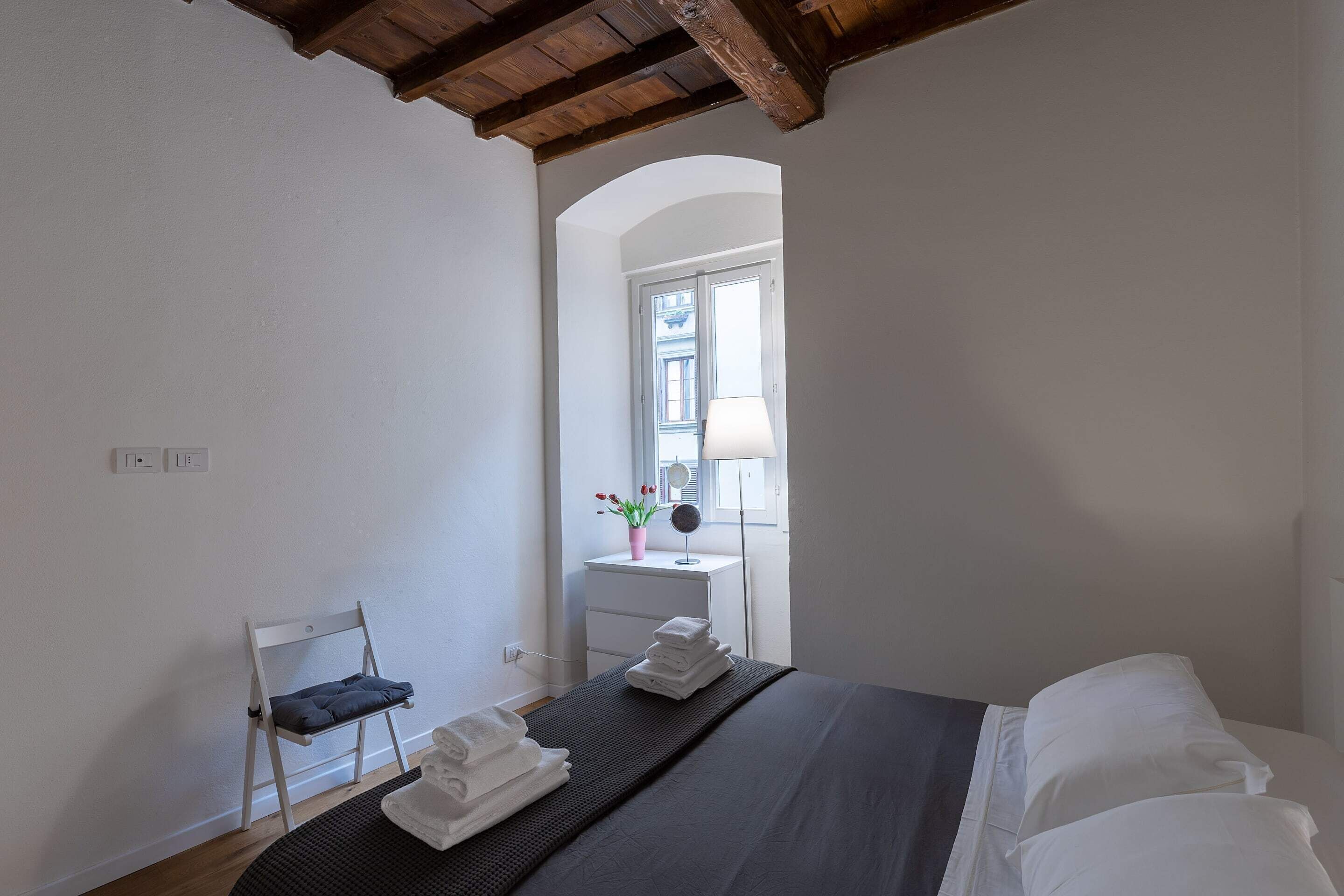 A bedroom with a bed, a chair, a small table with a lamp, and a window with a view.