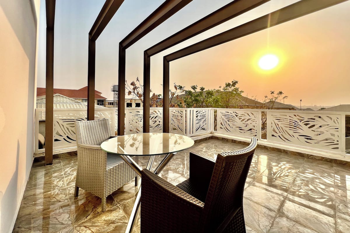 Un balcon avec une table en verre et des chaises en osier, donnant sur un quartier résidentiel au coucher du soleil.