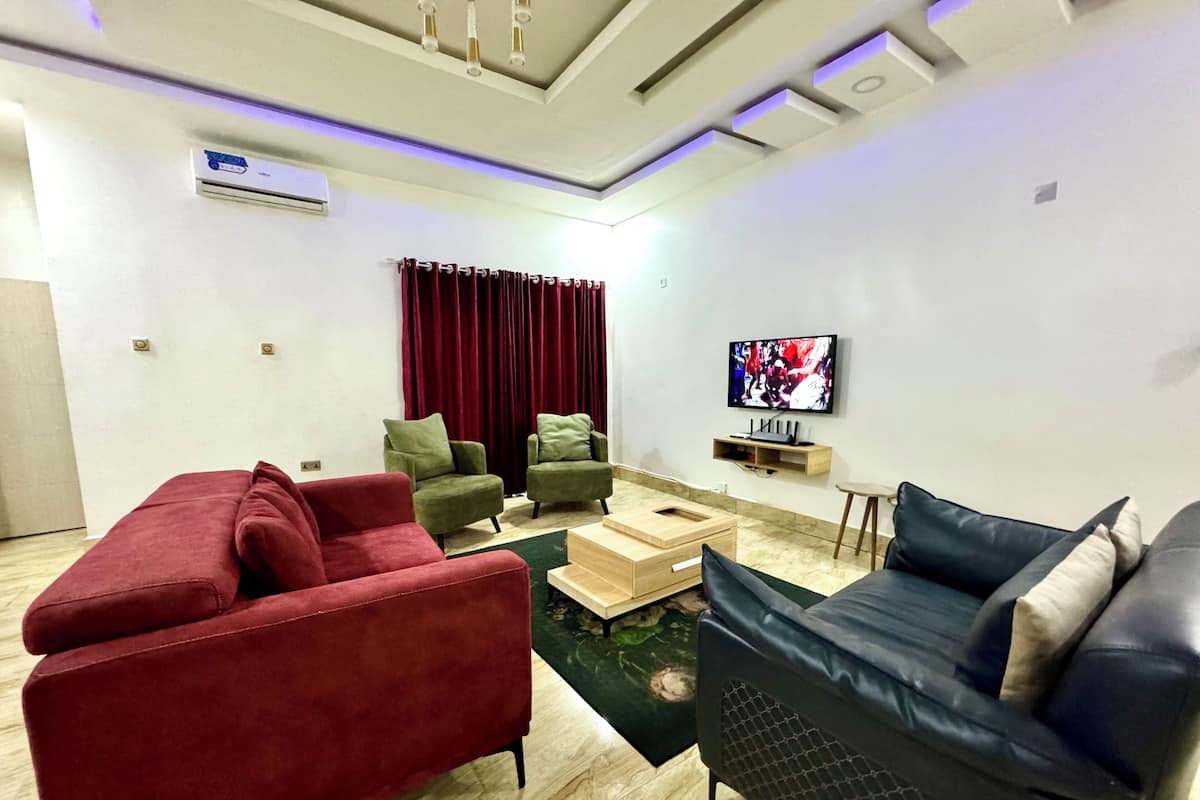 Un salon moderne avec un canapé rouge, des fauteuils verts, une table basse en bois et un canapé noir. Un téléviseur à écran plat est fixé au mur, et un luminaire est suspendu au plafond.