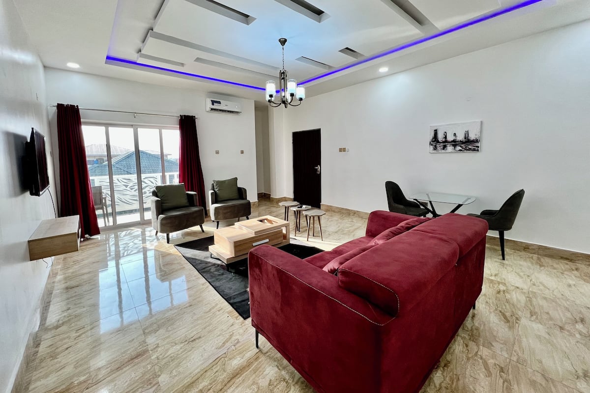 Un salon moderne avec un canapé rouge, deux fauteuils, une table basse en verre et un coin repas comprenant une petite table et des chaises.