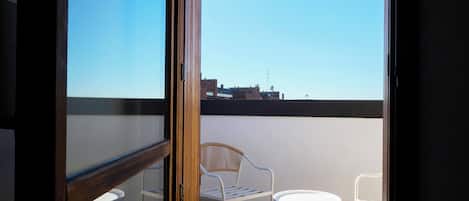 A balcony with two white chairs and a small table, a wooden door, and a clear view of buildings in the distance.
