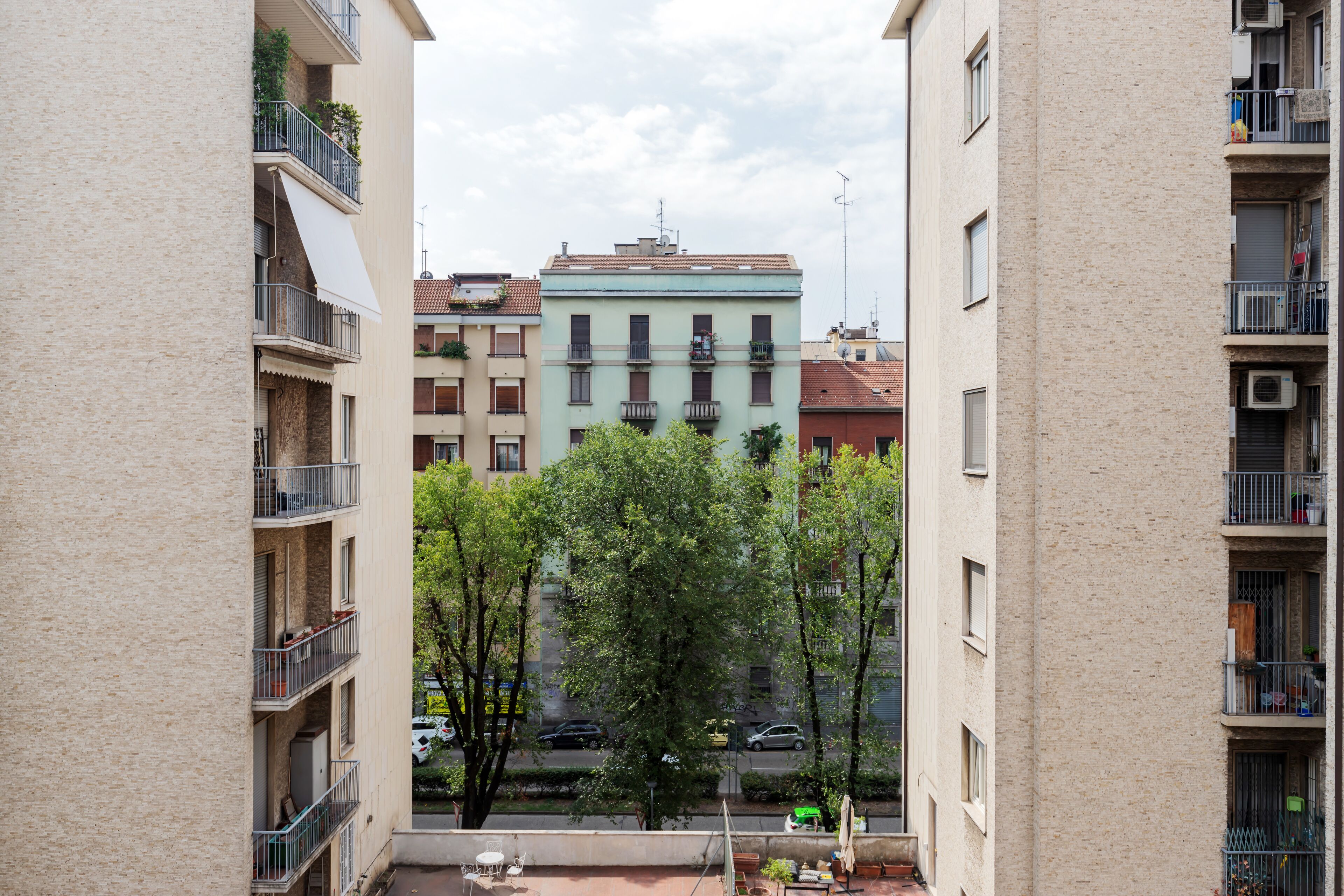 Une vue d’un immeuble résidentiel doté de nombreux balcons, avec une rue au premier plan et une rue bordée d’arbres en arrière-plan.