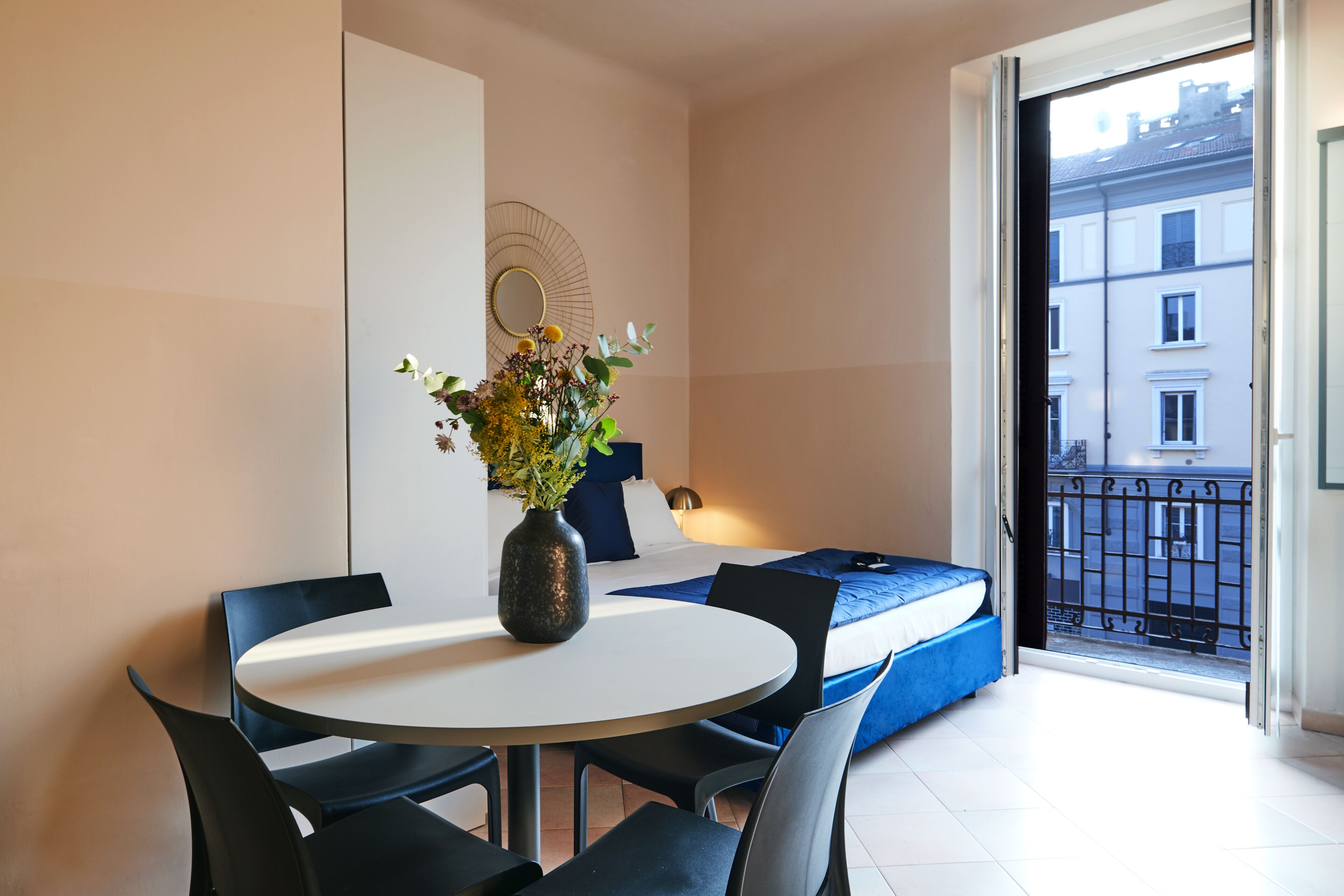 Een moderne hotelkamer met een ronde eettafel, twee stoelen, een bed met blauw beddengoed, een klein nachtkastje en een vaas met bloemen.