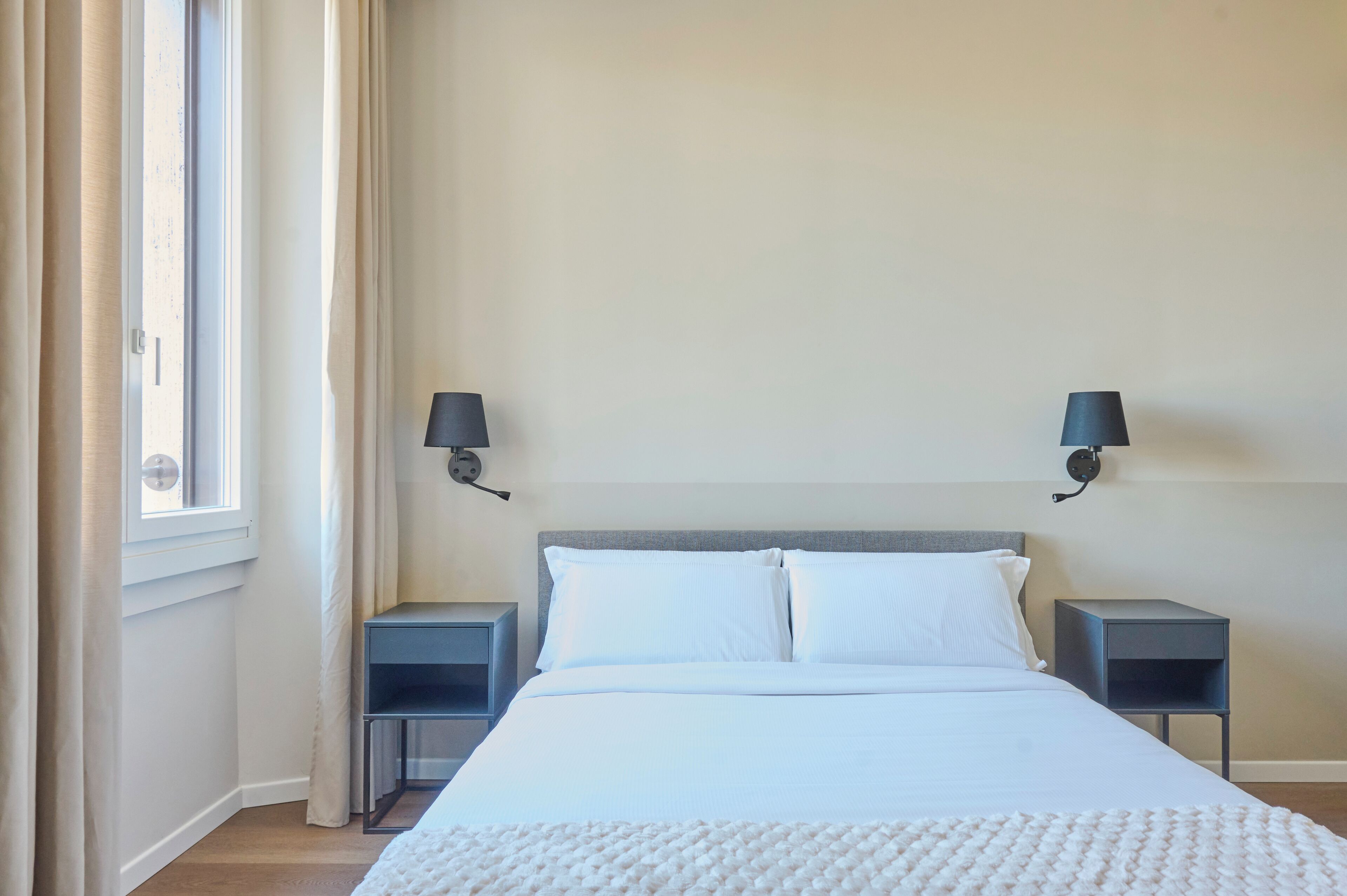 A bedroom with a bed, two bedside tables, a window with curtains, and wall-mounted lamps.