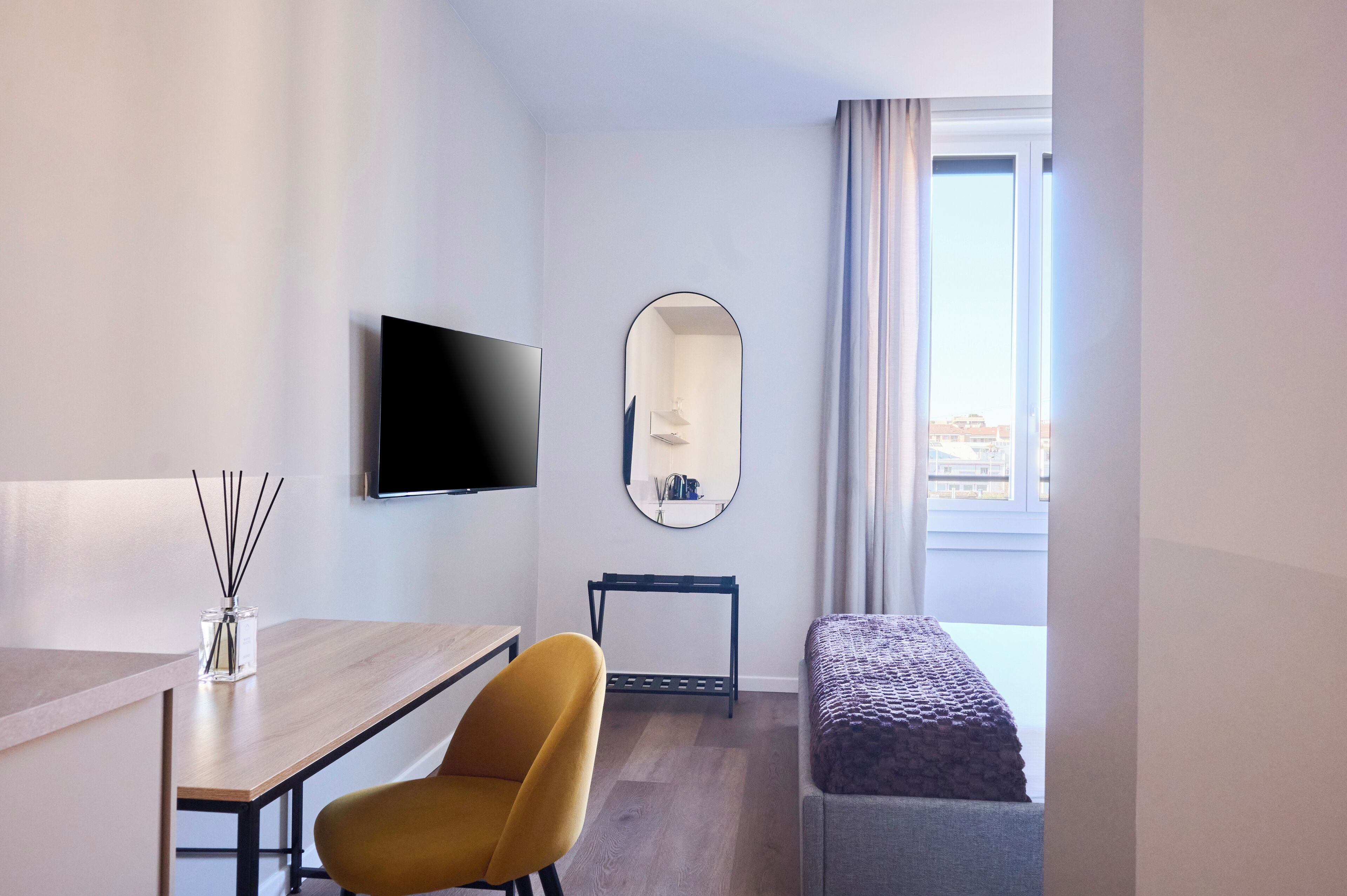 A hotel room with a flat-screen TV, a mirror, a wooden desk, a yellow chair, a bench, and a window with a view.