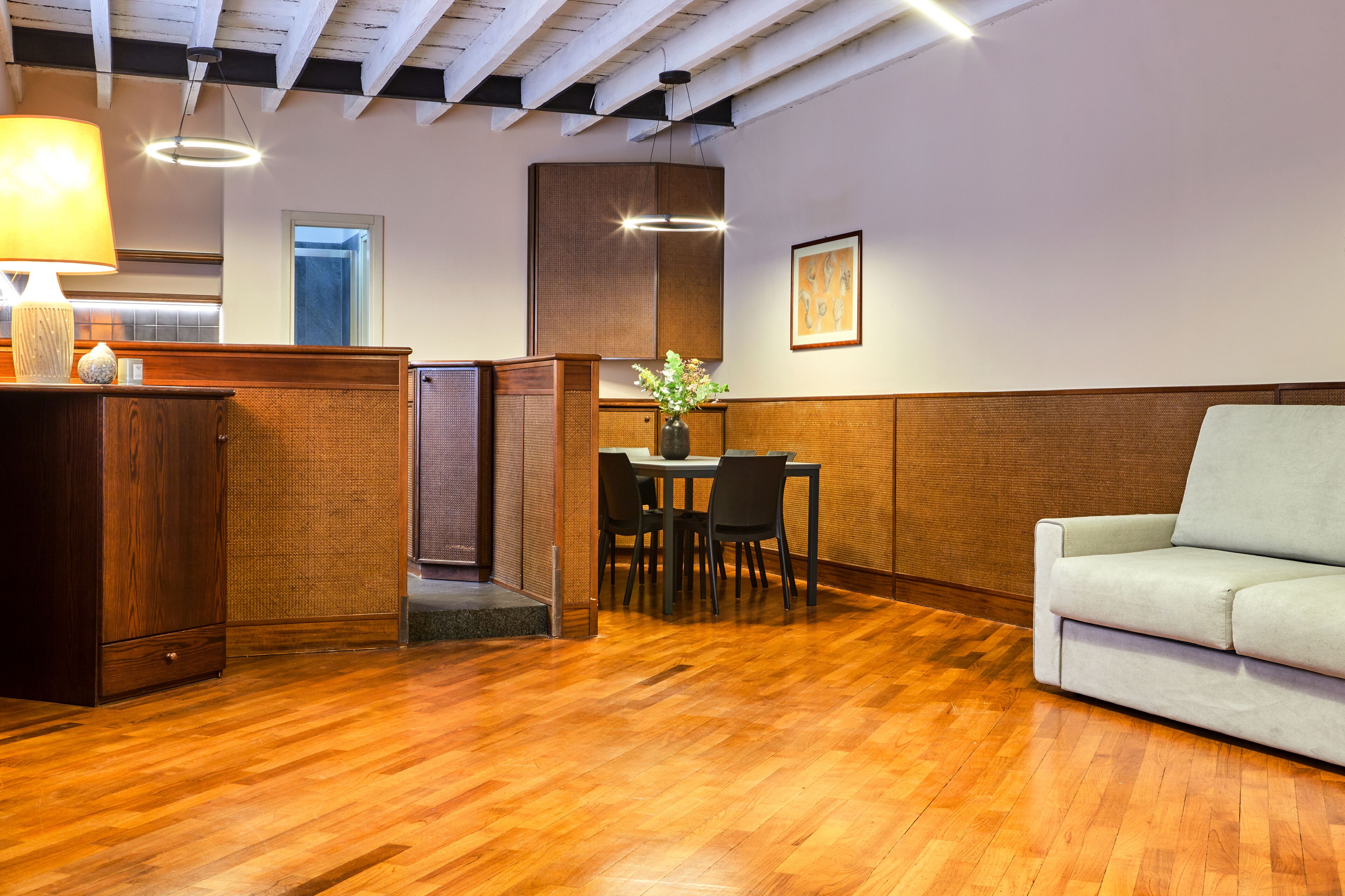 Een kamer met een houten vloer, een bank, een eettafel met stoelen, een lamp en een vaas met bloemen.