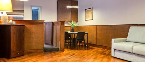 A room with a wooden floor, a sofa, a dining table with chairs, a lamp, and a vase with flowers.