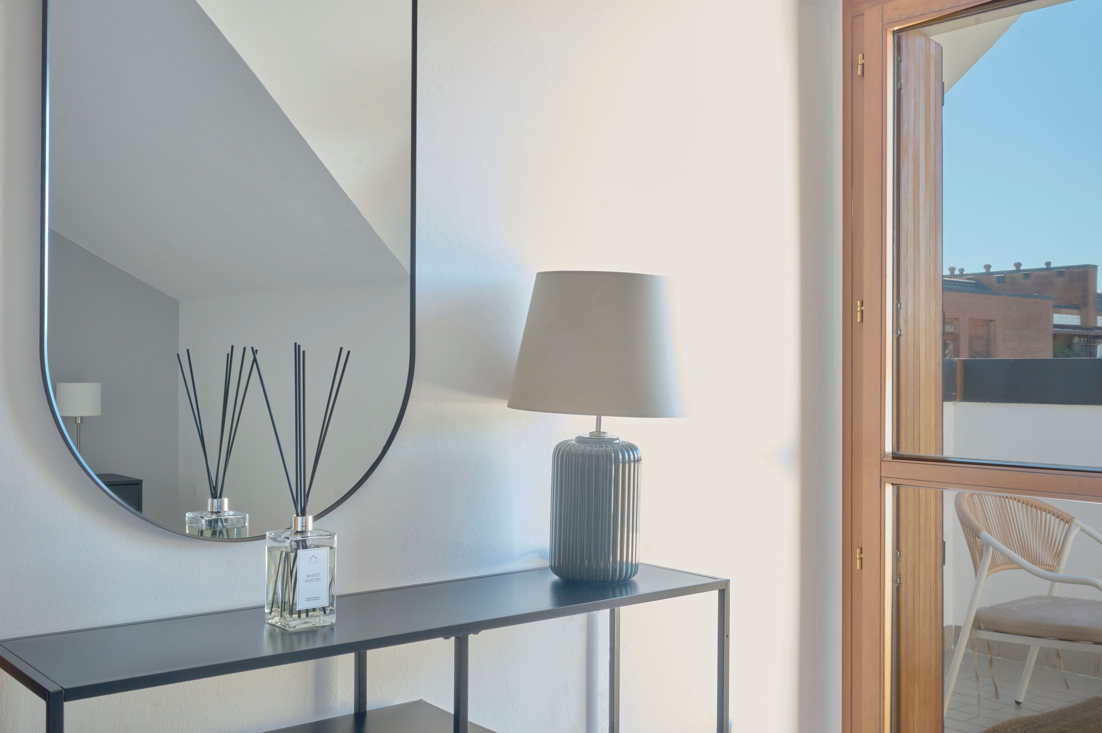 A modern room with a large mirror, a table with a lamp and perfume bottles, and a view of the outside.
