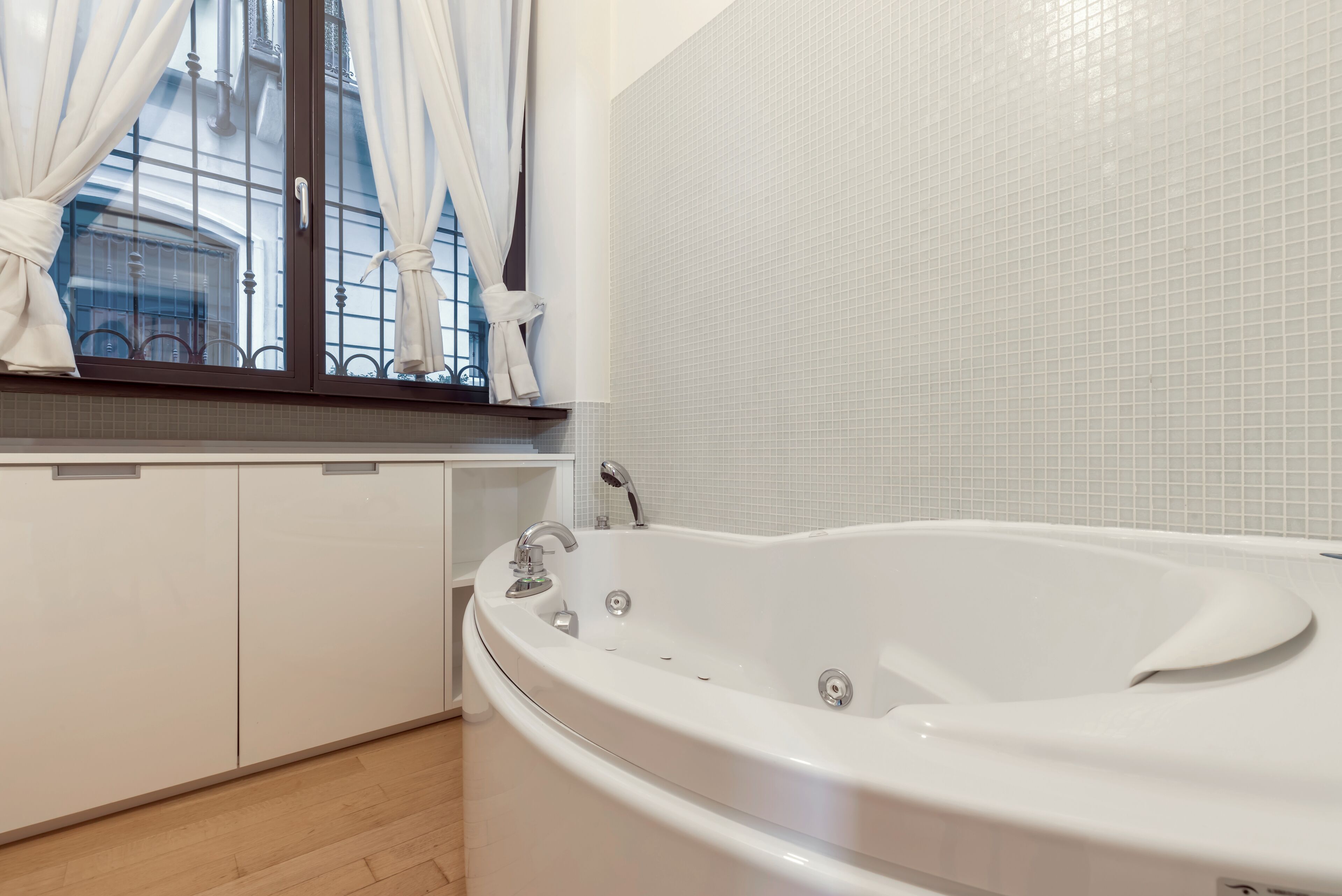 Un bagno con una vasca da bagno bianca, una finestra con tende e un muro rivestito di piastrelle.