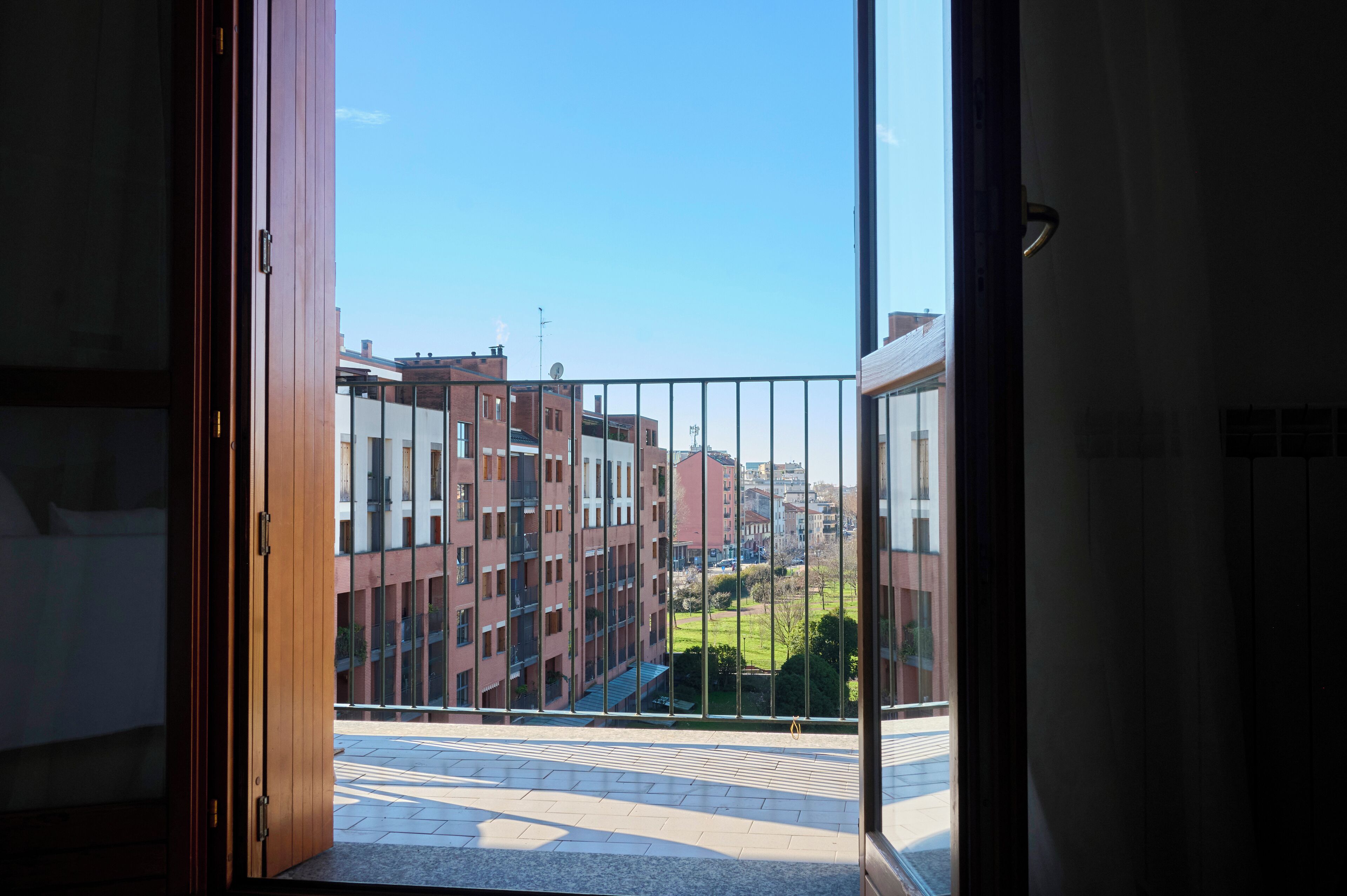 Une porte ouverte donnant sur un balcon avec vue sur un ensemble d’immeubles et un espace vert.