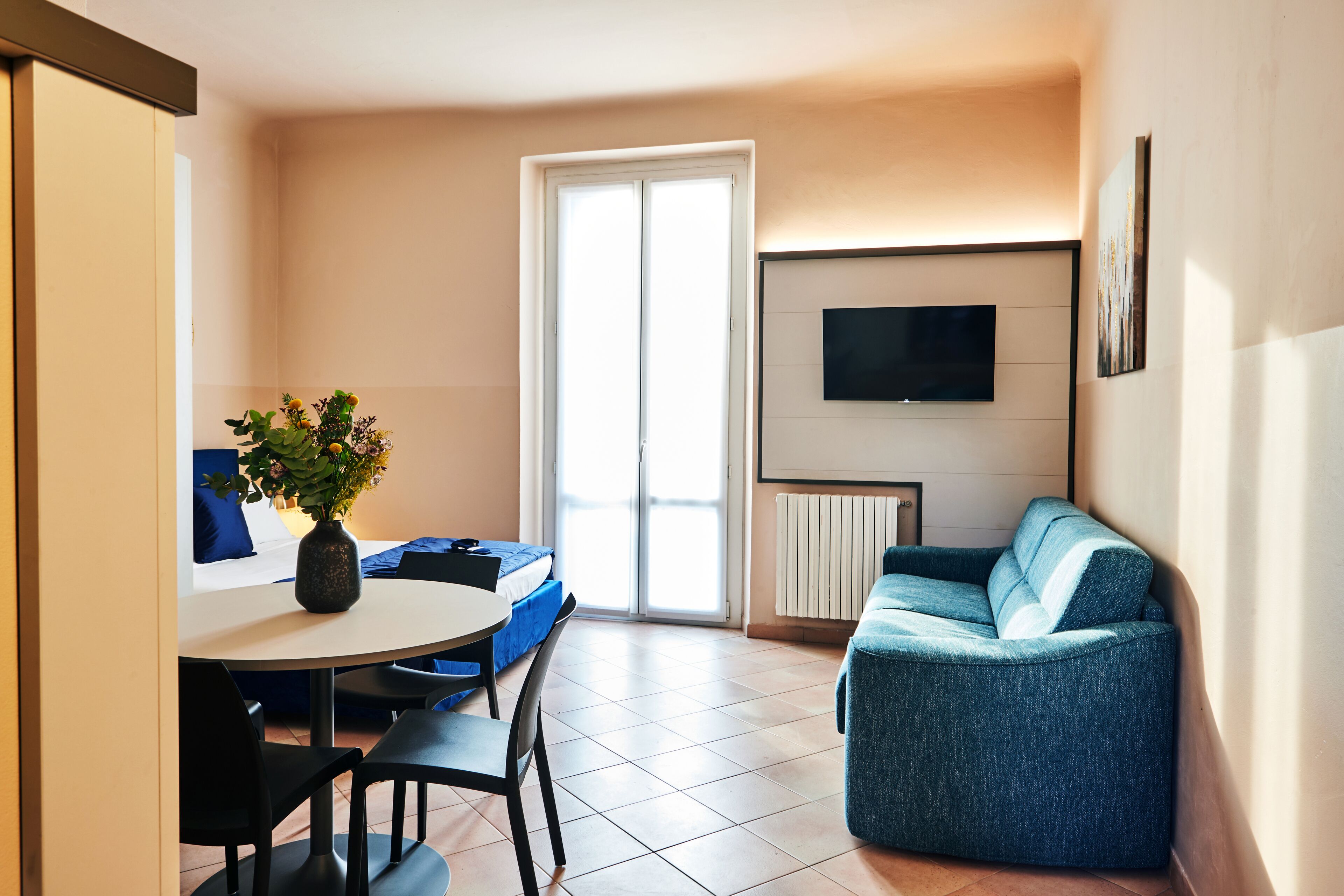 A compact living space with a dining area, a sofa, a TV mounted on the wall, and a sliding glass door leading to an outdoor area.