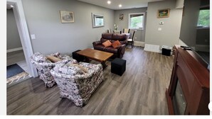 A spacious living room with a brown leather sofa, floral-patterned armchairs, a wooden coffee table, and a dining area with a table and chairs.