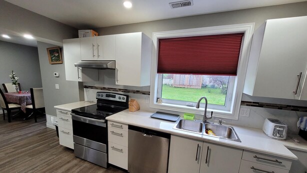 A modern kitchen with white cabinets, stainless steel appliances, a window with a red blind, and a view of a backyard.
