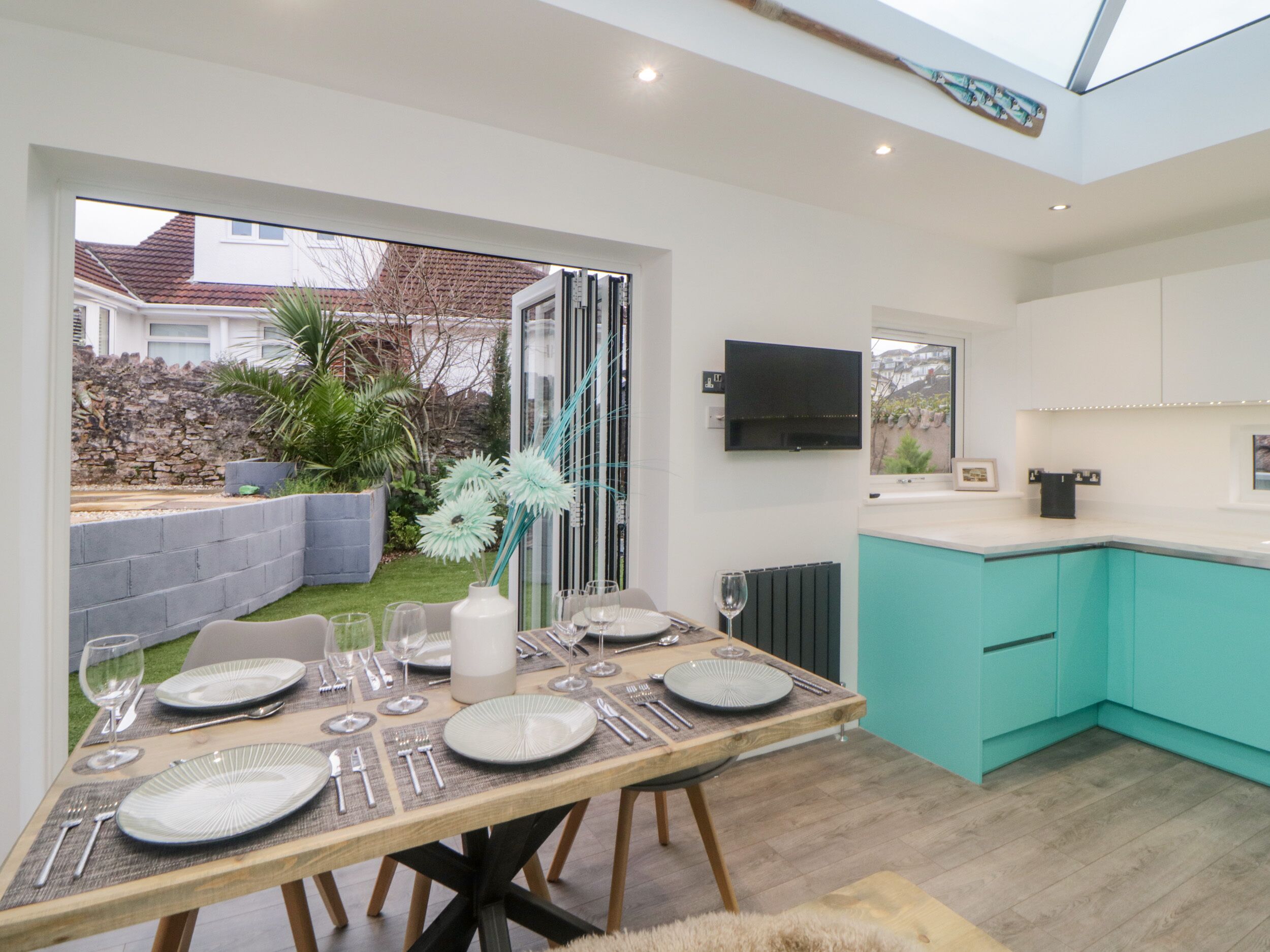 Una cocina moderna con armarios azul verdoso, una zona de comedor con mesa de madera dispuesta para seis personas, y vista al jardín a través de un ventanal.