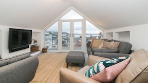 A spacious loft apartment with a large window, a flat-screen TV, and a cozy seating area with a sofa and ottoman. - Ingledene (Brixham)