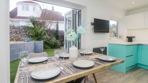 A dining area with a table set for four, a vase with flowers, and a view of a garden and house outside. - Ingledene (Brixham)
