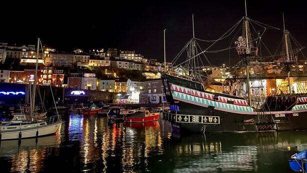 A harbor at night with boats docked, illuminated buildings in the background, and a prominent ship with colorful decorations. - Ingledene (Brixham)