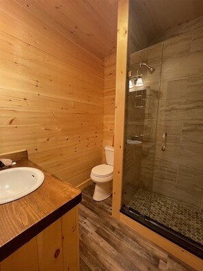 Ett badrum i en stuga med träpanel, en dusch med glasväggar, en vit handfat och en toalett.