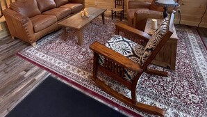 Interior de una cabaña de madera con escalera, sofá de cuero, mesa de centro, alfombra estampada y dos ventanas.