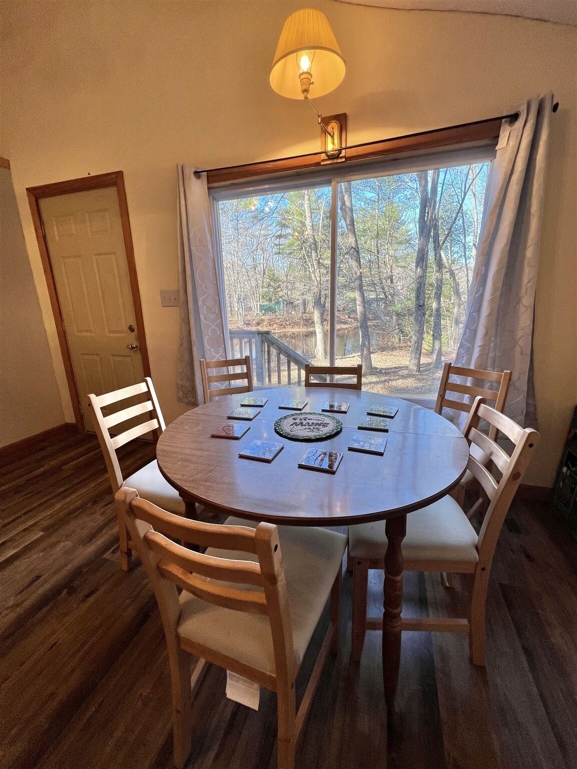 A round wooden dining table with six chairs, a lamp above, and a view of trees through a window.