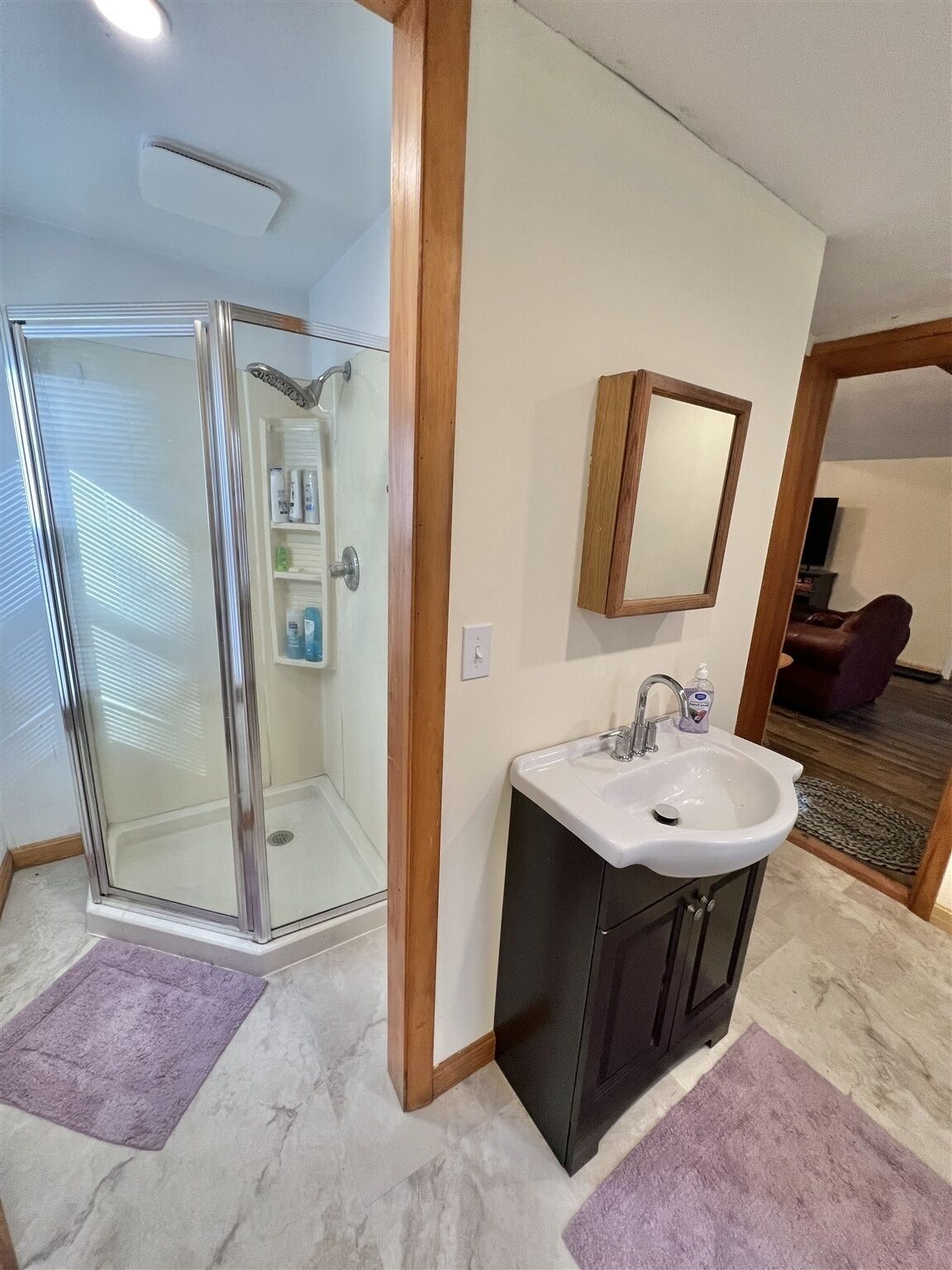 A bathroom with a glass shower enclosure, a sink with a mirror above it, and a purple rug on the floor.