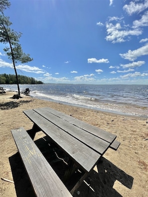 Un banco de madera en una playa arenosa, con cielo despejado y mar tranquilo al fondo.