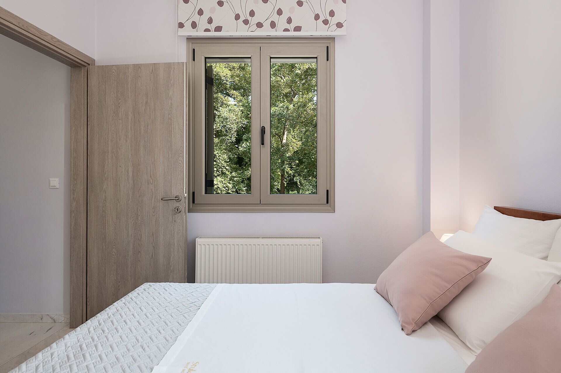 Une chambre à coucher avec un lit, un radiateur, une fenêtre donnant sur des arbres et une porte.