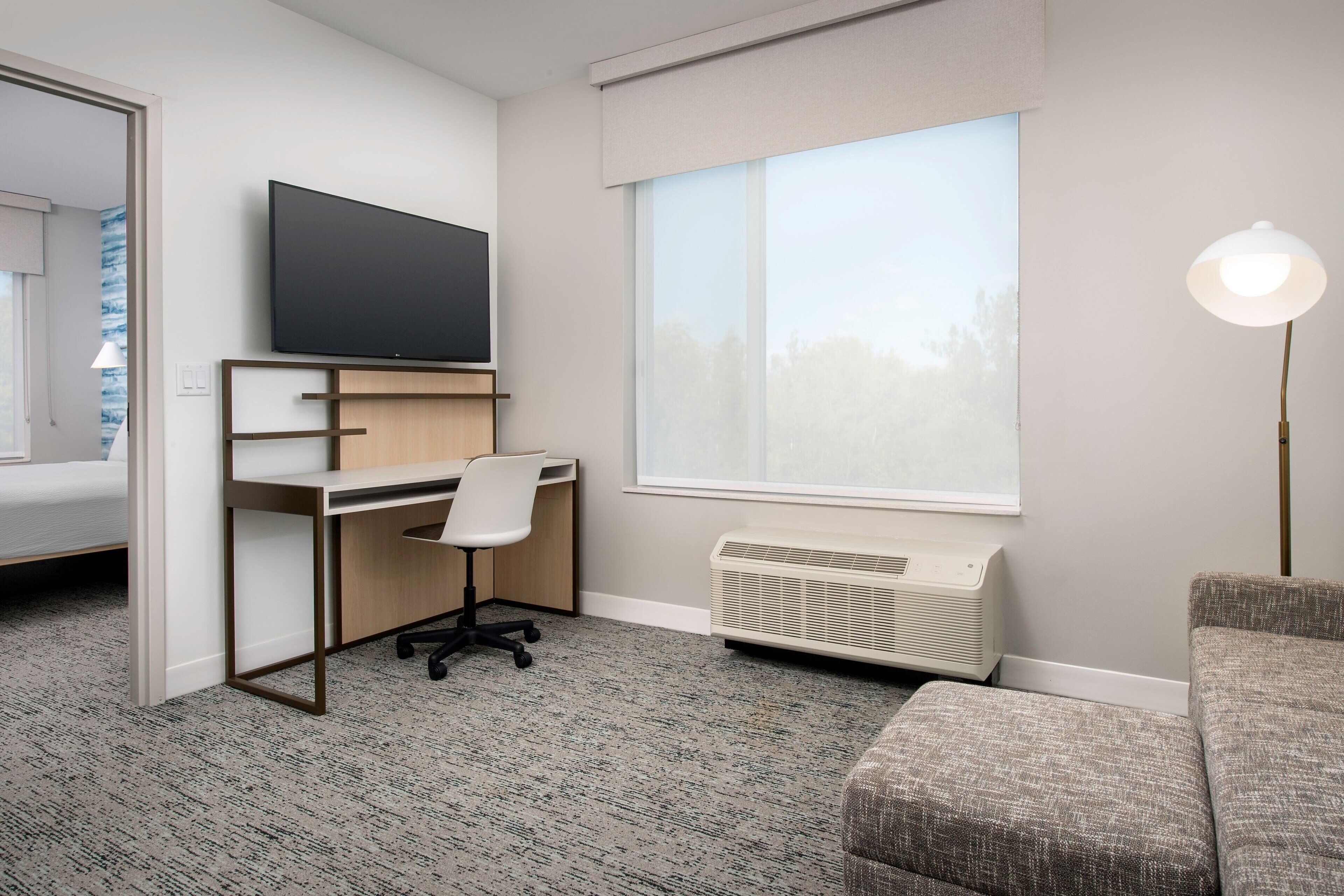 A hotel room with a bed, a desk with a chair, a TV, a window, and a lamp.