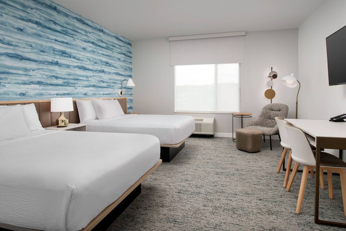 A hotel room with two beds, a desk, a chair, a lamp, and a large window.
