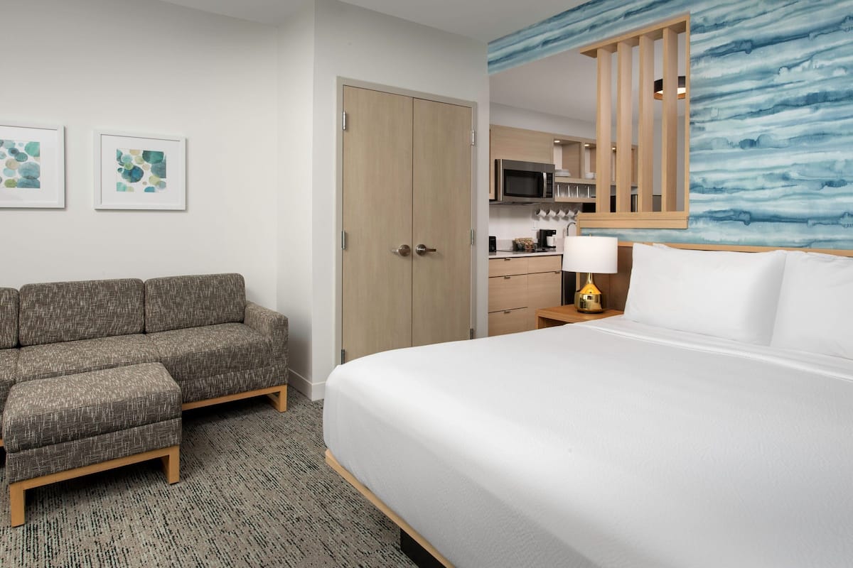 A hotel room with a bed, a sofa, a kitchenette, and artwork on the wall.