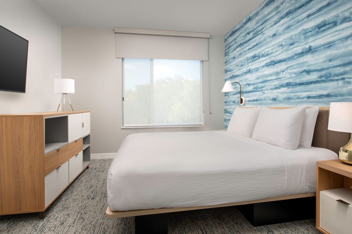 A modern hotel room with a large bed, a wooden nightstand, a desk with a lamp, a flat-screen TV, and a window with a view of trees.