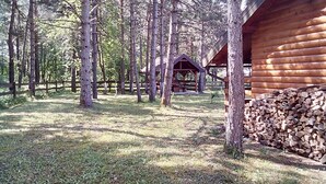 Une cabane en rondins dotée d’une terrasse en bois et d’un petit porche, entourée de grands arbres et d’une zone herbeuse.