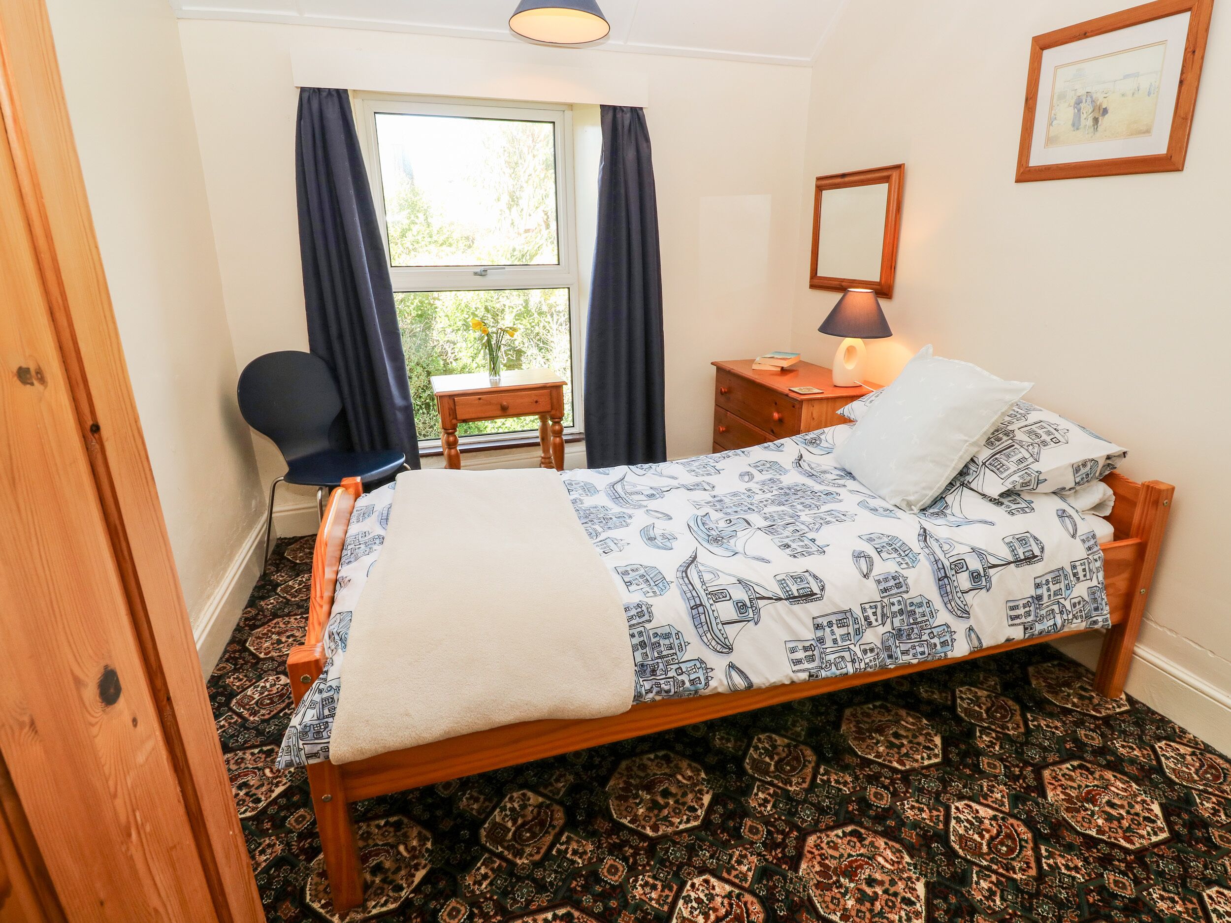 Un dormitorio ordenado con una cama, una silla, una mesita de noche, una lámpara y un ventanal con cortinas.