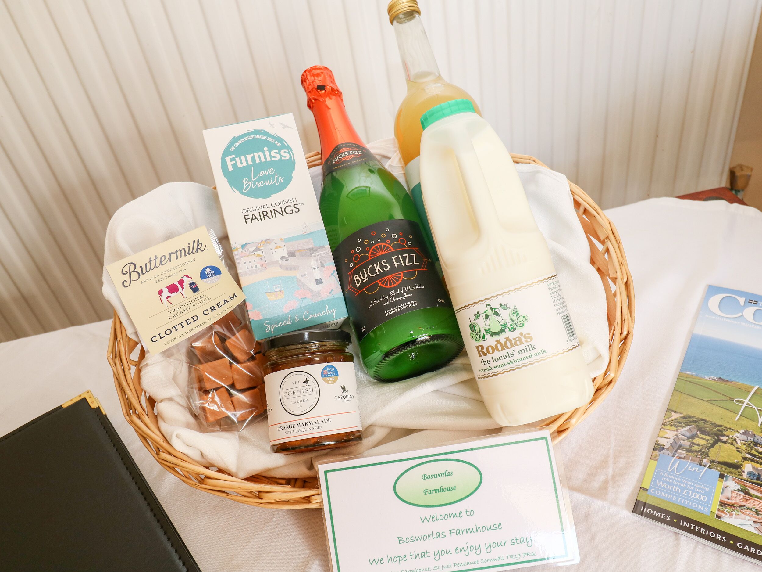 Un panier contenant divers produits alimentaires et une bouteille de vin, accompagné d’une pancarte de bienvenue pour la ferme Boswolas.