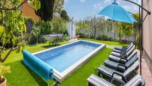 Une piscine entourée de verdure, avec des chaises longues et un parasol, sous un ciel dégagé.